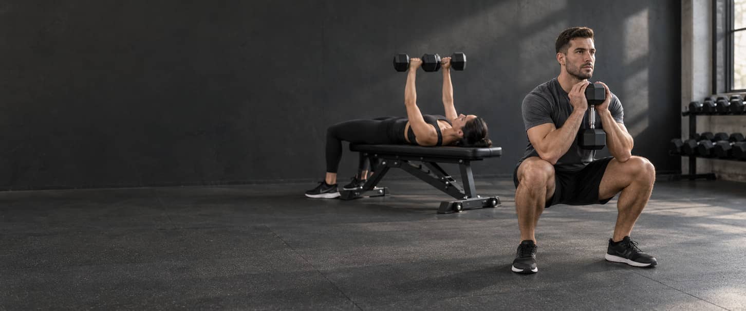 Man performing a dumbbell goblet squat with a woman doing a dumbbell bench press behind him